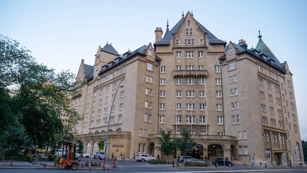 Fairmont Hotel Macdonald forward-facing view in Edmonton The Fairmont Hotel Macdonald stands tall with its château-style turrets and stone façade in this forward-facing view from across the street, highlighting the grand entrance, tree-lined sidewalk and elegant heritage architecture that makes it Edmonton’s iconic “castle on the hill.”