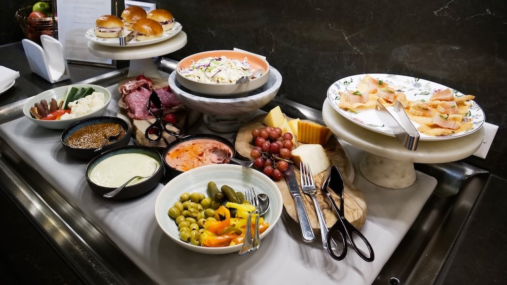 Evening canapé spread at Fairmont Gold Lounge An inviting evening canapé spread at the Fairmont Hotel Macdonald’s Gold Lounge displays sliders, cured meats, smoked fish, cheeses, olives, dips, vegetables and fresh fruit arranged on marble stands, showcasing the generous and beautifully presented selection available to guests.