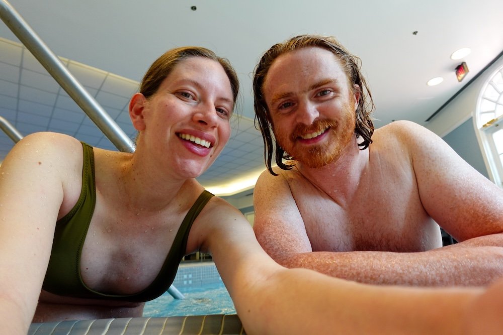 Samuel and Audrey relaxing at the Fairmont Hotel Macdonald spa pool Nomadic Samuel Jeffery and Audrey Bergner of That Backpacker smile together at the edge of the Fairmont Hotel Macdonald’s indoor spa pool, enjoying a warm and relaxing babymoon moment with gentle lighting and calm water after exploring Edmonton’s historic château hotel.