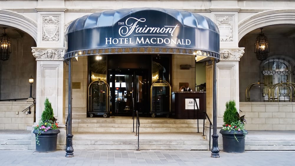Main entrance of Fairmont Hotel Macdonald in Edmonton, Alberta, Canada with historic stone architecture Main entrance of Fairmont Hotel Macdonald in Edmonton, Alberta, Canada, with its branded canopy, stone arches, luggage carts, flower planters and hotel doorway.