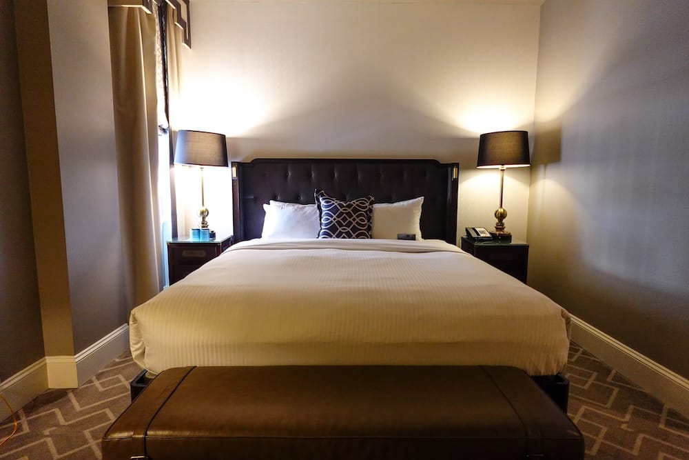Cama king de habitación Fairmont Gold en el Hotel Macdonald de Edmonton durante nuestra babymoon de una noche Cama king en nuestra habitación Fairmont Gold del Fairmont Hotel Macdonald en Edmonton, Alberta, Canadá, con sábanas blancas, cabecera capitoné oscura, lámparas a juego, ventana angosta y banco de cuero.