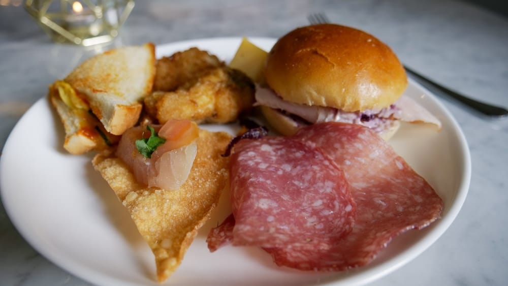 Canapés de la tarde del Fairmont Gold Lounge, Fairmont Hotel Macdonald, Edmonton, Alberta, Canadá Un plato blanco de canapés de la tarde del Fairmont Gold Lounge en el Fairmont Hotel Macdonald en Edmonton, Alberta, Canadá, que incluye salame cortado, una minihamburguesa, wantanes fritos y crostini, apoyado sobre una mesa de mármol.