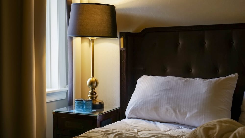 Detalle de habitación Fairmont Gold con lámpara y ventana angosta en el Hotel Macdonald de Edmonton Detalle junto a la cama de una habitación Fairmont Gold en el Fairmont Hotel Macdonald de Edmonton, Alberta, Canadá, con cabecera capitoné oscura, ropa de cama blanca, lámpara de bronce, ventana pequeña y vasos azules.
