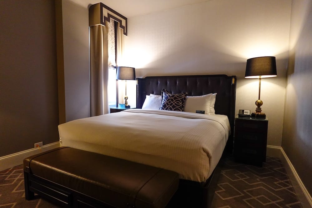 Guest room at Fairmont Hotel Macdonald in Edmonton, Alberta, Canada with king bed and classic interior details Guest room at Fairmont Hotel Macdonald in Edmonton, Alberta, Canada, with a large bed, dark upholstered headboard, bedside lamps, patterned carpet and a narrow window.