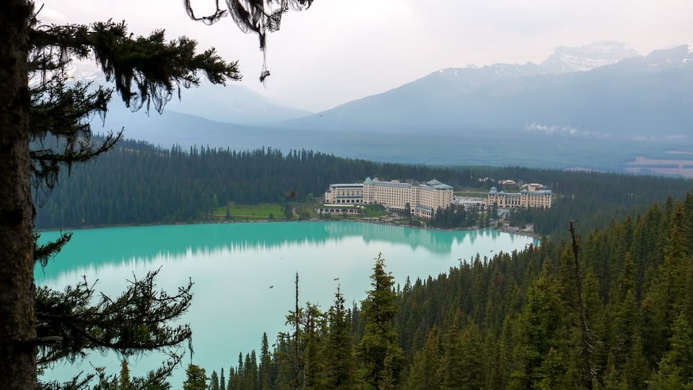 Vista elevada del agua turquesa de Lake Louise desde Fairview Lookout en Banff National Park, Alberta, Canadá, con laderas boscosas, picos montañosos y el Fairmont Chateau Lake Louise al otro lado del agua.