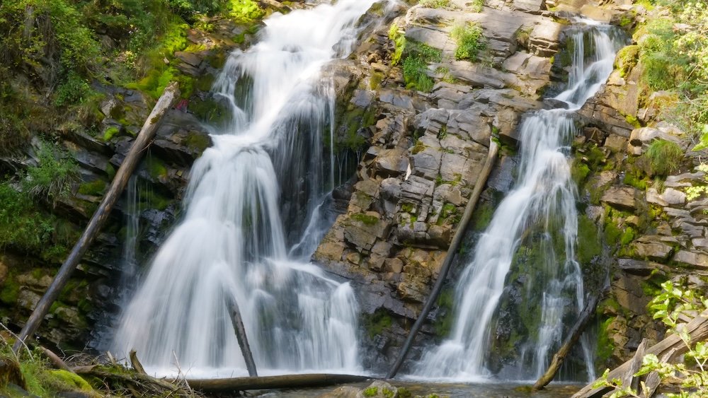 Fairy Creek Falls in Fernie, British Columbia captured with silky motion-blur water cascading over layered rock ledges, framed by mossy stone, fallen logs, and lush forest greenery—one of the most photogenic, easy waterfall stops near town.