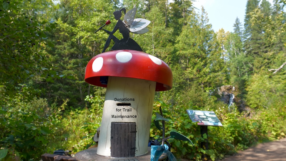 Trail maintenance donation box shaped like a red mushroom beside the Fairy Creek Falls trail in Fernie, British Columbia, encouraging hikers to support upkeep of this family-friendly forest hike near the waterfall trailhead area popular with visitors.
