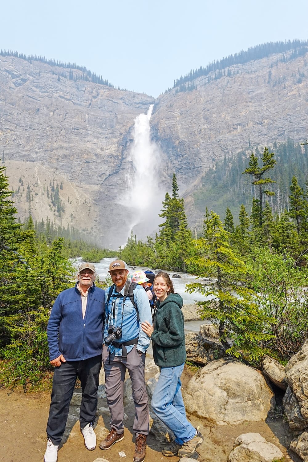 Daniel Bergner, Samuel Jeffery, and Audrey Bergner smile together beside the Kicking Horse River at Takakkaw Falls in Yoho National Park, British Columbia, with Aurelia in a carrier and the misty waterfall filling the valley behind them.