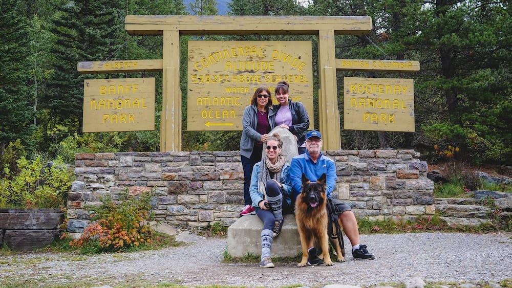 Family group at the Continental Divide sign, Banff and Kootenay National Parks, Alberta An extended family group sits and stands with their German Shepherd Togo at the Continental Divide sign, with the Banff National Park and Kootenay National Park boundary markers behind them.
