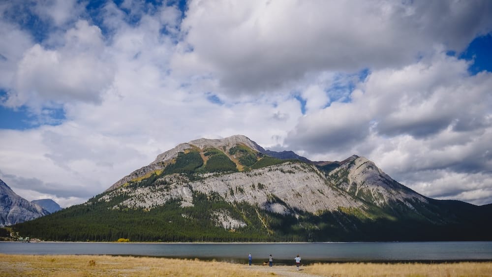 Family at Lower Kananaskis Lake, Peter Lougheed Provincial Park, Alberta Small figures stand along the shoreline of Lower Kananaskis Lake in Peter Lougheed Provincial Park, Alberta, dwarfed by a massive mountain wall rising directly behind the water.