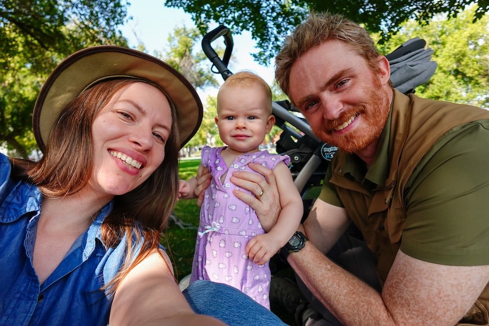 Family park day in Cranbrook with Nomadic Samuel and Audrey Bergner Nomadic Samuel Jeffery, Audrey Bergner of That Backpacker, and their daughter Aurelia smiling together during a sunny family outing in Cranbrook, British Columbia. They sit under leafy trees at a quiet park, taking a relaxed break during their East Kootenay travels and filming their Cranbrook travel video.