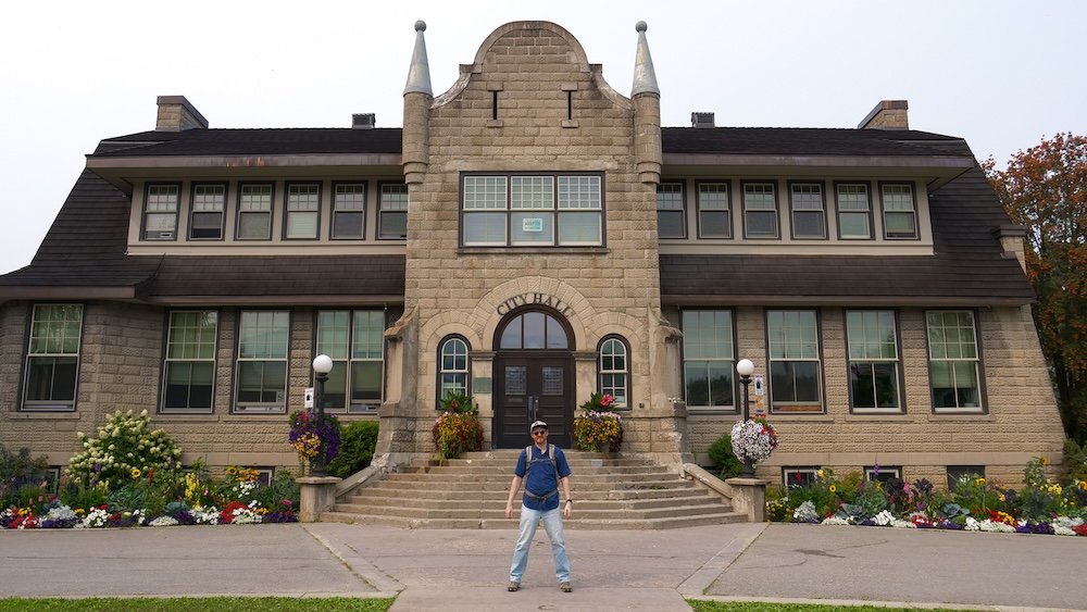 Fernie City Hall gardens with Nomadic Samuel Jeffery Fernie, British Columbia City Hall with colorful flower beds as Nomadic Samuel Jeffery stands on the front walkway, framed by stone-and-brick heritage architecture and twin turrets—an easy downtown stop on the heritage walk.