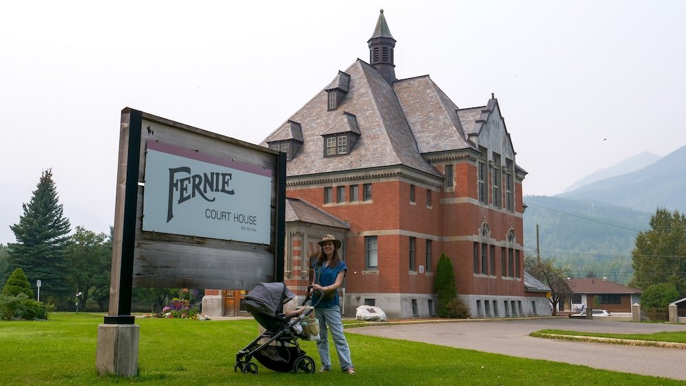 Fernie, British Columbia courthouse scene as Audrey Bergner pushes baby Aurelia in a stroller beside the Fernie Court House sign, with the historic red-brick building and hazy mountain backdrop showing the town’s heritage vibe.
