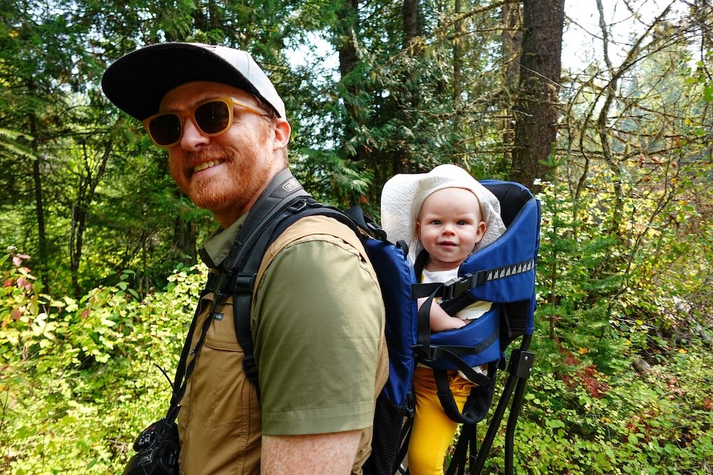 Fernie, British Columbia forest hiking scene as Nomadic Samuel hikes the Fairy Creek Falls trail while carrying baby Aurelia in a backpack carrier, enjoying a family-friendly adventure through lush greenery and working up a big appetite after exploring Fernie’s mountain trails.
