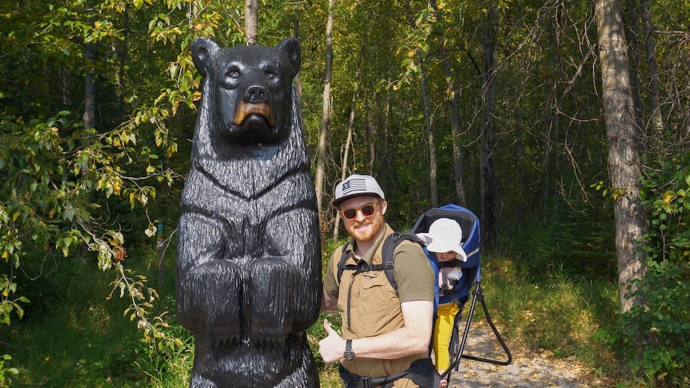 Fairy Creek Falls near Fernie, BC shows Nomadic Samuel hiking beside a carved bear statue while carrying baby Aurelia in a backpack carrier, highlighting an easy forest trail that makes this scenic waterfall a popular family-friendly day trip from Cranbrook.
