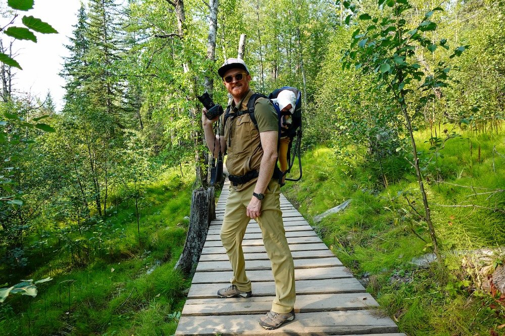 Fernie, British Columbia forest boardwalk on the Fairy Creek Falls trail as Nomadic Samuel hikes through lush greenery while carrying baby Aurelia in a backpack carrier, capturing a family-friendly hiking moment in the mountains near Fernie.