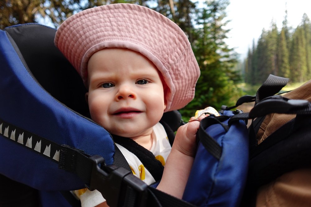 Fernie, British Columbia family hiking moment showing baby Aurelia smiling comfortably in a backpack carrier on a forest trail, surrounded by soft greenery and dappled light—highlighting how Fernie’s easy hikes work well for traveling with a baby.