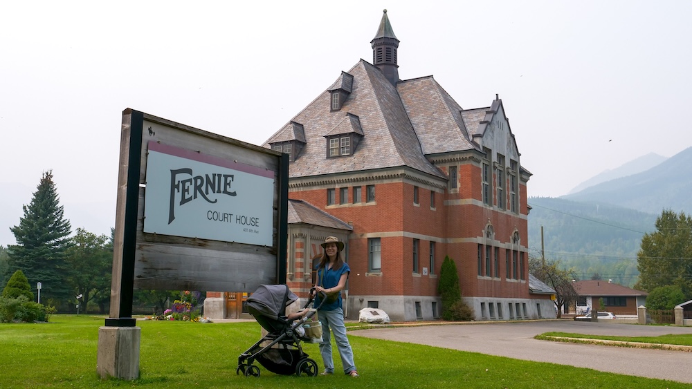 Fernie, British Columbia heritage scene at the historic Fernie Courthouse, with Audrey Bergner pushing baby Aurelia in a stroller on green lawn, showcasing small-town charm and mountain scenery in the Canadian Rockies