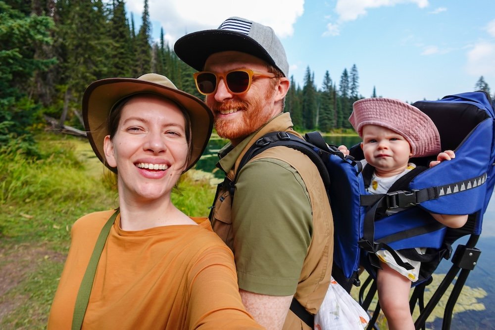 Island Lake Lodge family hike selfie in Fernie, BC Fernie, British Columbia selfie at Island Lake Lodge: Nomadic Samuel Jeffery and Audrey Bergner hike with baby Aurelia in a backpack carrier beside an alpine lake, with evergreen forest and peaks in the background.