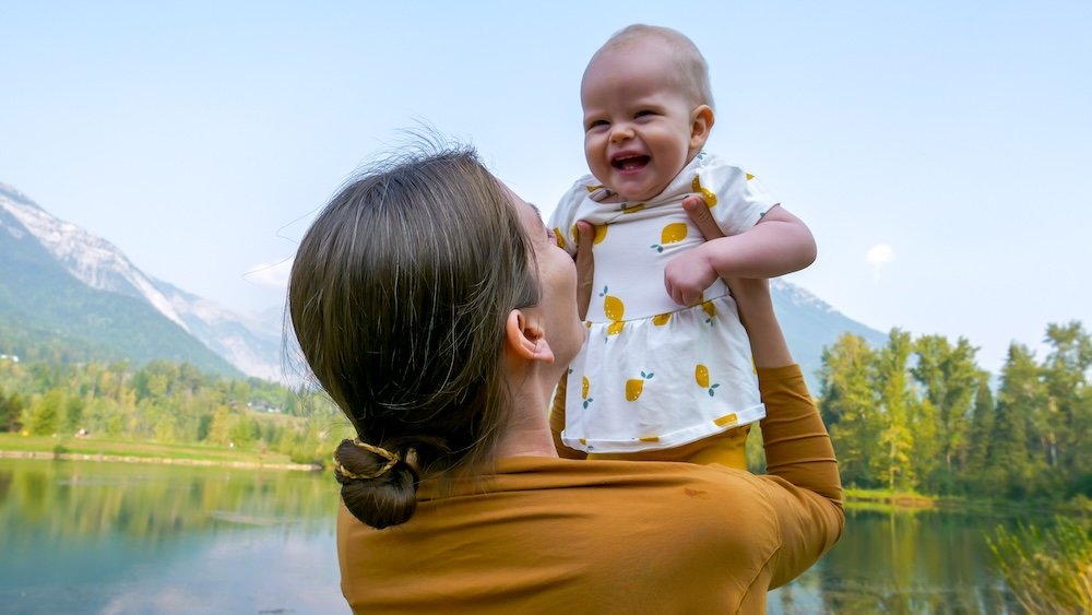 Fernie, British Columbia lakeside scene at Maiden Lake as Audrey Bergner lifts baby Aurelia while enjoying mountain views, capturing a joyful family travel moment that highlights how calm, scenic spaces in Fernie are perfect for visiting with young children.