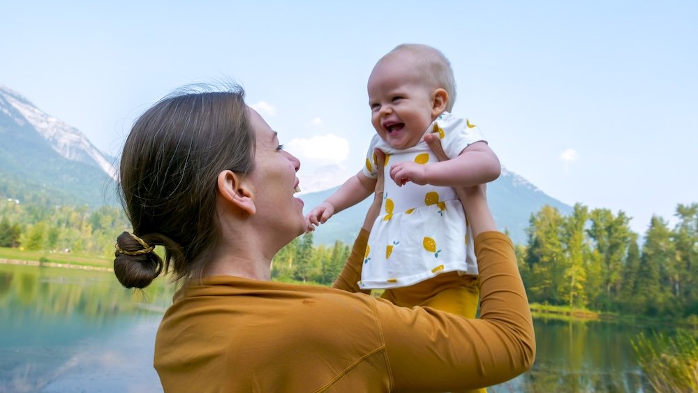 Mother and baby enjoying Maiden Lake in Fernie, BC Fernie, British Columbia moment at Maiden Lake as Audrey Bergner lifts baby Aurelia into the air, sharing a joyful laugh beside calm water with mountain peaks and forest behind them on a warm late-summer family visit.