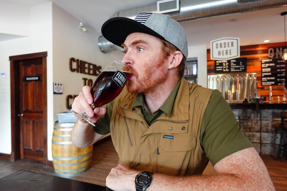 Nomadic Samuel enjoying a Ridgewalk Red at Fernie Brewing Company Nomadic Samuel Jeffery enjoys a cold pint of Ridgewalk Red Ale inside Fernie Brewing Company after a long hike, seated at a wooden table with the taproom’s warm lighting, rustic barrels, and “Cheers to Beers” signage creating a cozy après-adventure atmosphere.