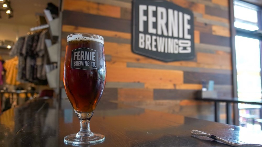 Ridgewalk Red Ale at Fernie Brewing Company A freshly poured Ridgewalk Red Ale sits on a polished wooden table inside Fernie Brewing Company’s taproom, with the brewery’s bold wall logo and warm wood-panel interior softly blurred in the background to highlight the rich amber beer as the star of the scene.