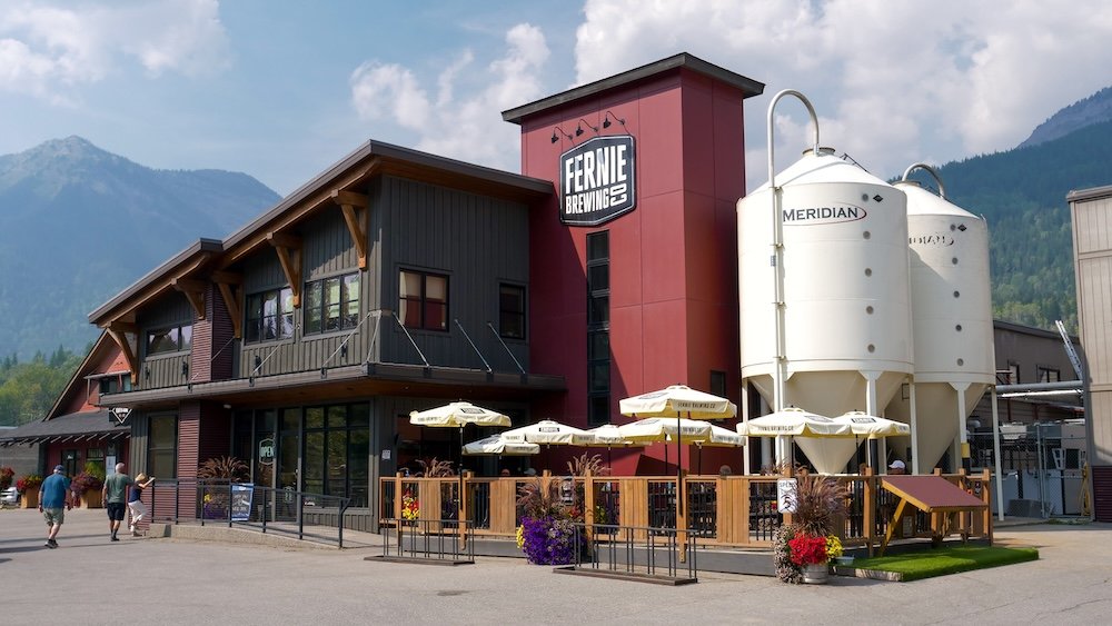 Fernie Brewing Company with mountain backdrop in Fernie, BC Fernie Brewing Company in Fernie, British Columbia, with its tasting room patio, outdoor umbrellas, and towering fermentation tanks set against a dramatic mountain backdrop, showing why this craft brewery is a popular post-hike stop for visitors exploring the Canadian Rockies.