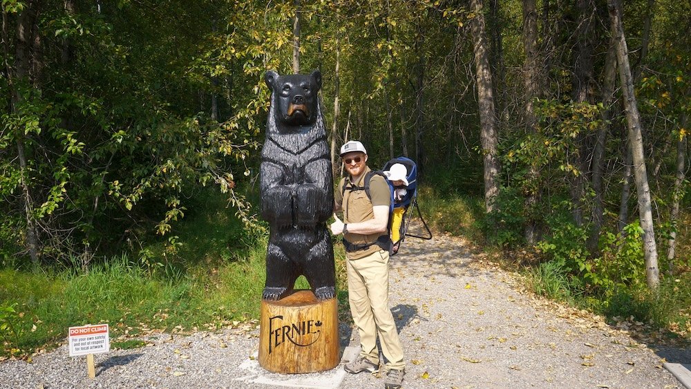 Fernie, British Columbia pre-hike stop with Nomadic Samuel and baby Aurelia posing beside the iconic bear sculpture on a forest trail, marking the playful start to a family-friendly outing before hiking to Fairy Creek Falls just outside town.
