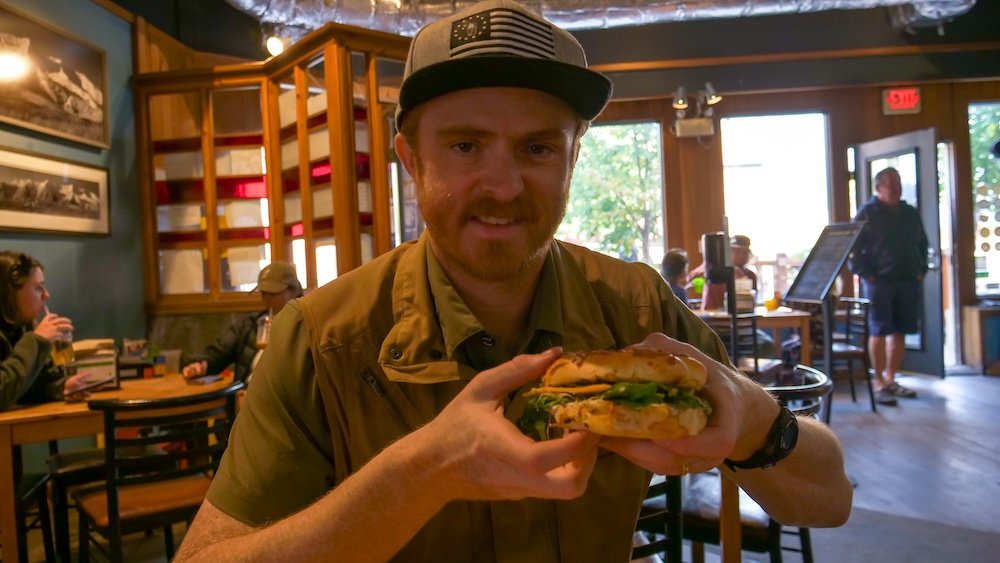 Fernie, British Columbia breakfast at Big Bang Bagels with Nomadic Samuel holding the Avolauncher bagel sandwich inside the shop, highlighting a popular local stop before hiking and exploring Fernie’s walkable downtown.