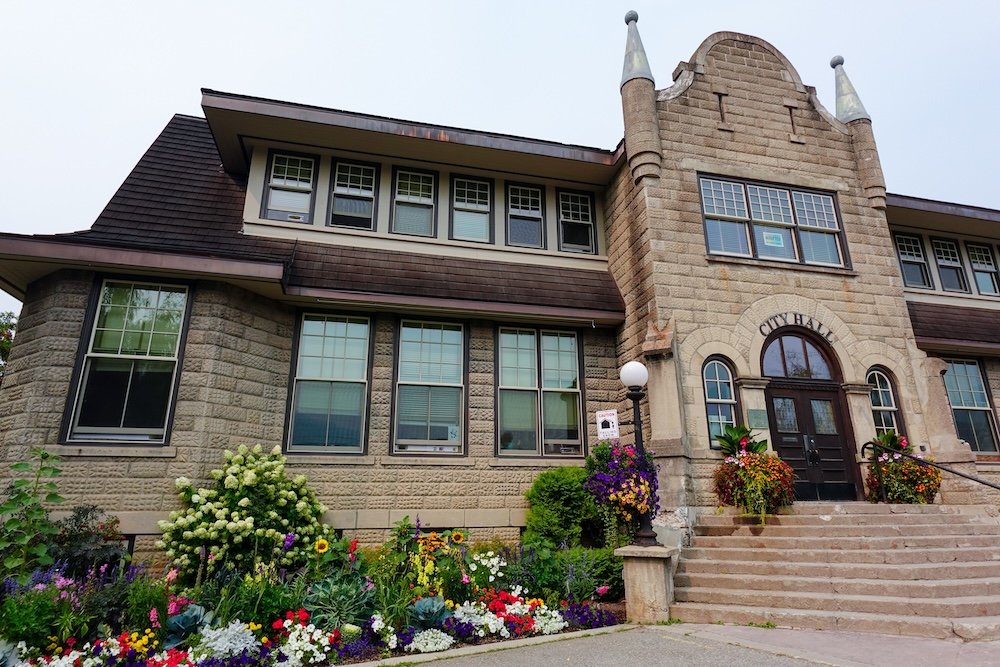 Fernie, British Columbia heritage architecture at City Hall, showing the stone-built historic building framed by colourful flower gardens along the Fernie Heritage Walk, highlighting the town’s walkable downtown and resilient brick-and-stone civic core.
