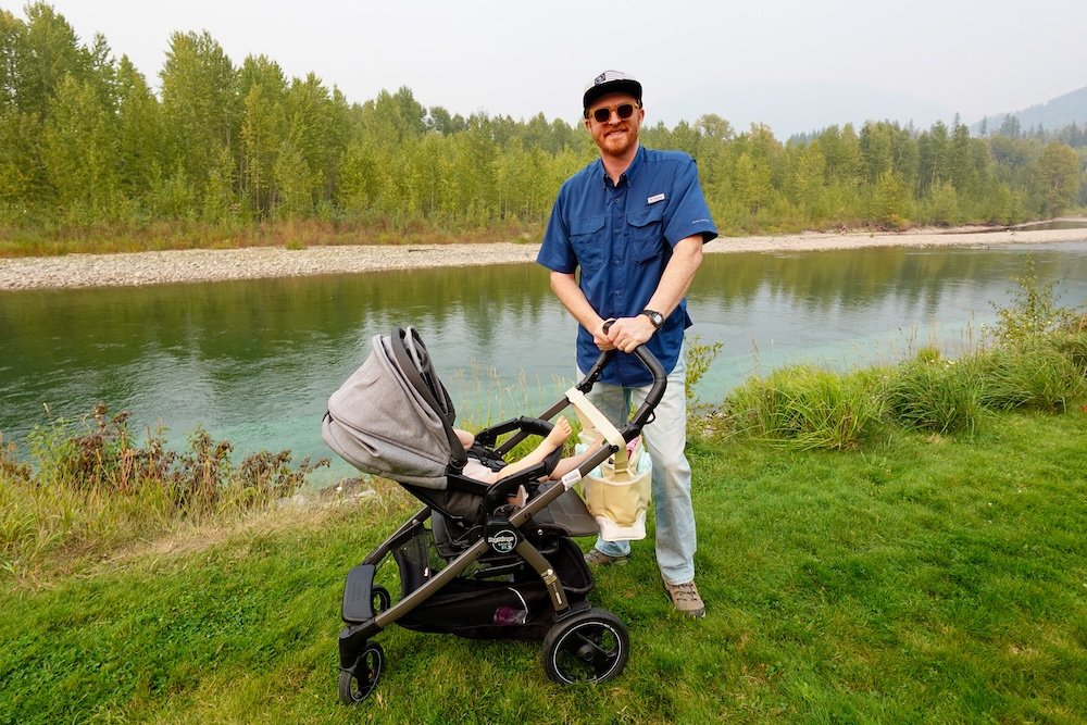 Fernie, British Columbia riverside scene showing Nomadic Samuel walking with a stroller beside a calm river and forested banks, highlighting how easy it is to enjoy nature, fresh air, and family-friendly outdoor spaces on a relaxed day trip to Fernie.