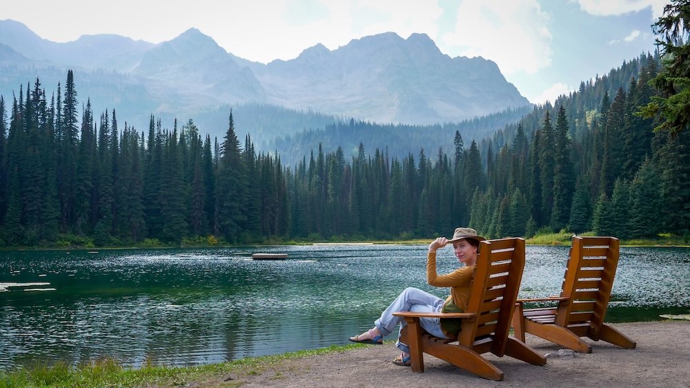Fernie, British Columbia lakeside views at Island Lake Lodge with Audrey Bergner of That Backpacker relaxing in a wooden chair beside the water, framed by evergreen forest and dramatic mountain peaks on one of Fernie’s most scenic and tranquil easy outings.
