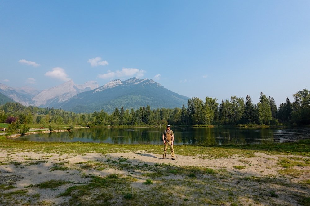 Fernie, British Columbia views at Maiden Lake showing Nomadic Samuel standing small against the shoreline, calm reflective water, and towering mountain backdrop, emphasizing the scale of Fernie’s scenery on an easy lakeside walk popular with families and beginners.
