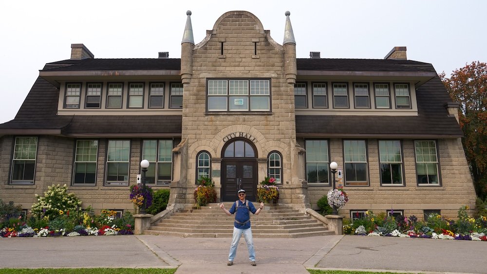 Nomadic Samuel at Fernie City Hall Nomadic Samuel standing in front of Fernie City Hall in British Columbia, framed by colorful flower beds and historic stone architecture, capturing a cheerful stop on our walking exploration of downtown Fernie’s heritage core.
