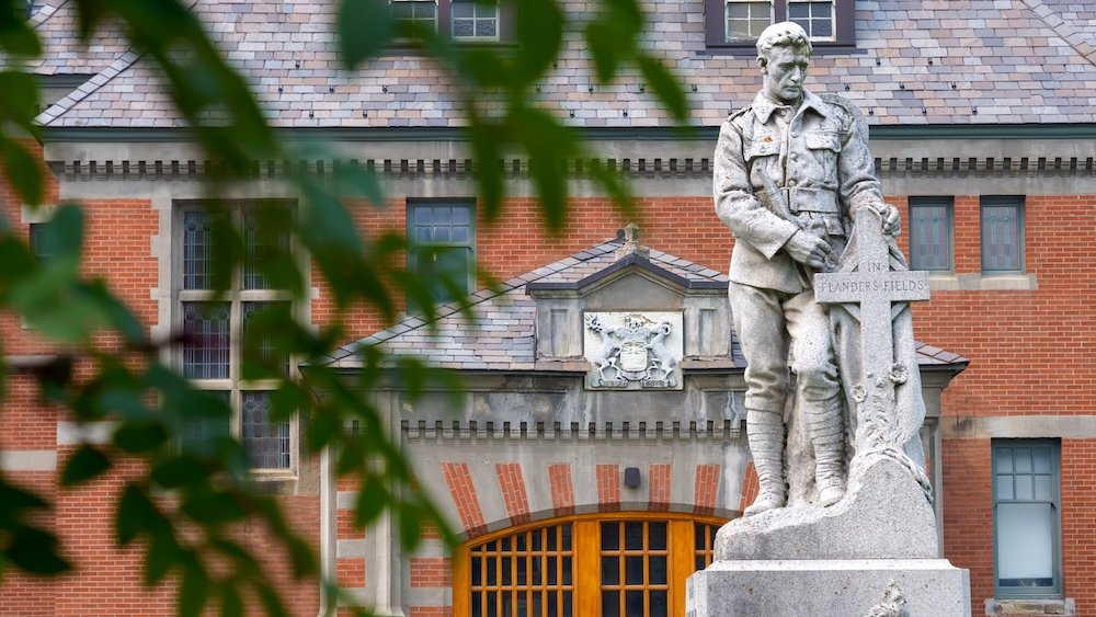 Fernie Courthouse in British Columbia with a stone war memorial statue framed through soft green leaves, set against historic brick architecture and arched windows—an artistic, heritage-rich downtown Fernie scene blending history, texture, and place.