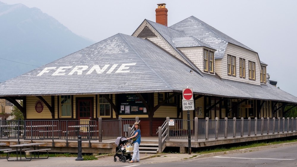 Audrey Bergner pushes a stroller with baby Aurelia outside Fernie’s historic CPR Station, now known as the Arts Station, during a self-guided heritage walking tour, with the wooden railway building and mountains creating a classic Fernie backdrop.
