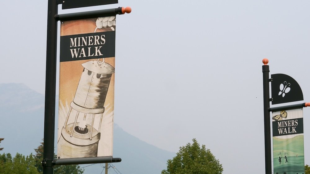 Miner’s Walk heritage route in Fernie Miner’s Walk street banners in Fernie, British Columbia, marking the historic walking route that interprets the town’s coal-mining past, labour history, and rebuilding era, with mountain views faintly visible beyond downtown.
