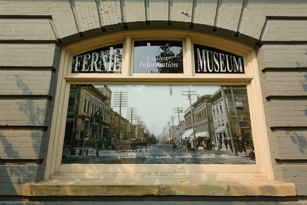 Fernie Museum visitor information window set into a historic brick building, featuring an archival street photograph that shows downtown Fernie in the early 1900s, offering context and orientation before starting a self-guided heritage walking tour.
