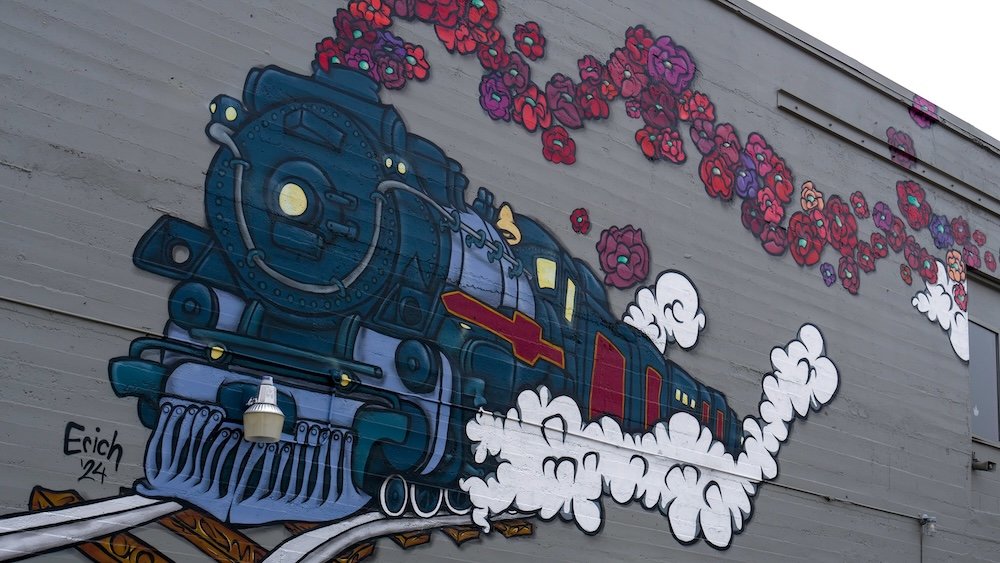 A colorful mural in downtown Fernie depicts a vintage steam train bursting through clouds of white steam and red poppies, symbolizing the town’s railway and mining heritage, painted on a large exterior wall along the self-guided heritage walking tour route in Fernie, British Columbia.
