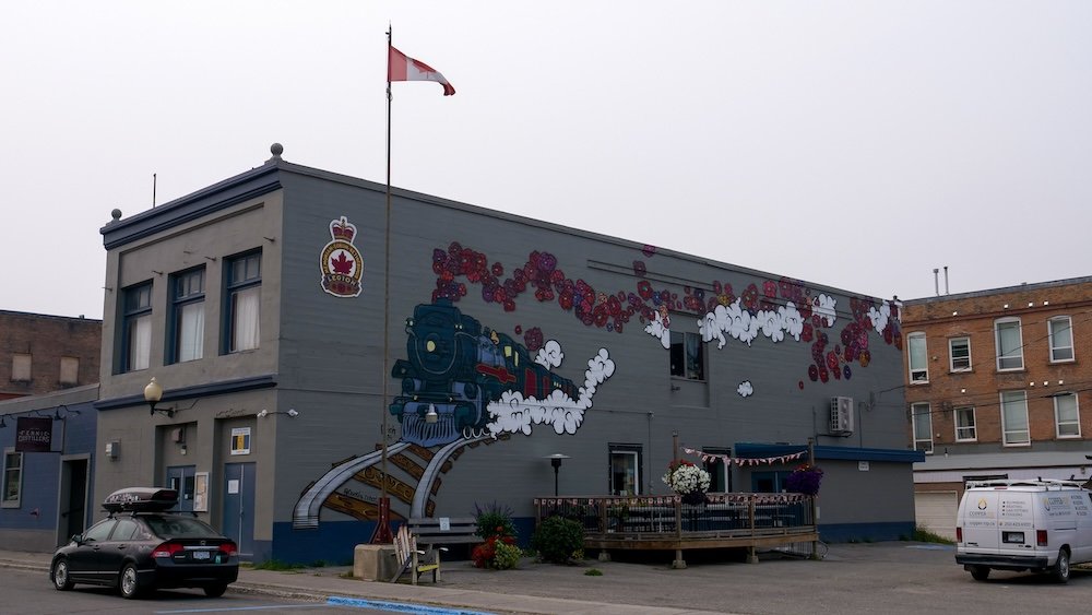 Fernie downtown train mural with Canadian flag A colourful train mural painted on a downtown Fernie building, featuring a steaming locomotive, red floral details, and a Canadian flag flying above, reflecting Fernie’s railway roots and small-town heritage in British Columbia.