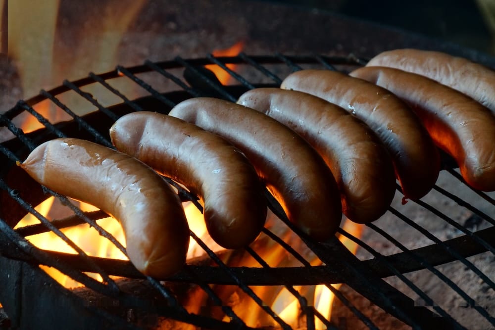 Finnish sausages cooking on a metal grill above an open campfire during an outdoor meal in Nuuksio National Park, showing a simple Finnish forest food tradition near Helsinki.
