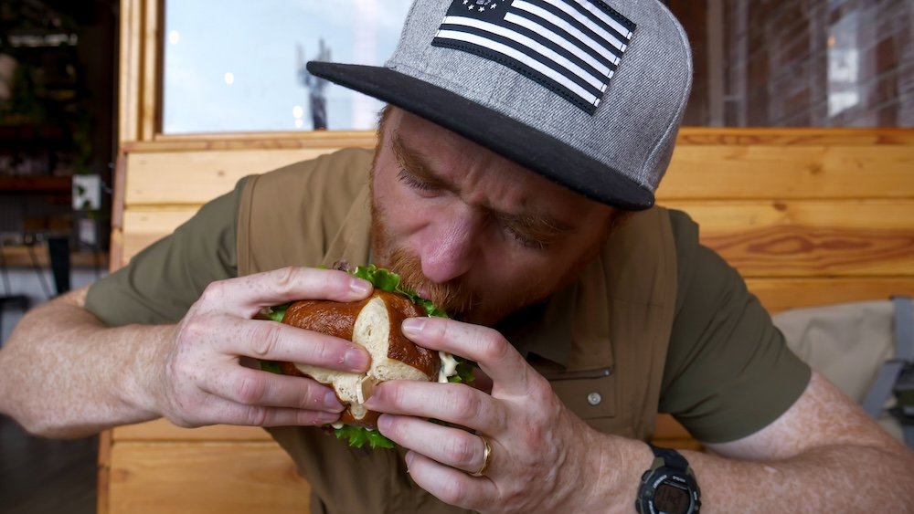 Fire Hall Kitchen & Tap in Cranbrook, British Columbia, with Nomadic Samuel taking a big bite of a legendary gourmet burger inside the converted fire station restaurant, highlighting one of the most memorable and satisfying food stops on a Cranbrook day trip.
