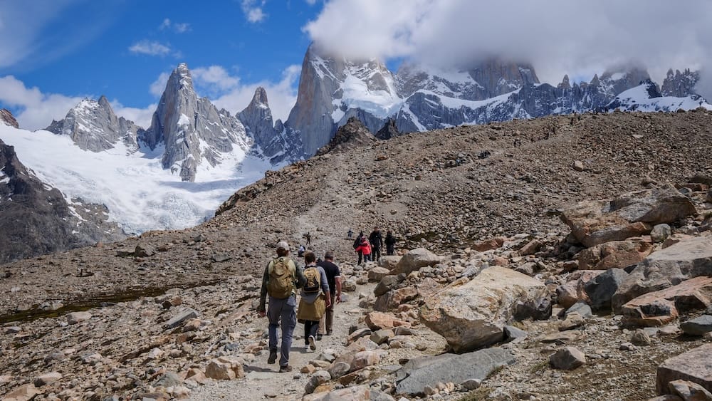 Fitz Roy Moraine Trail, El Chaltén — Santa Cruz, Patagonia, Argentina | Trekkers with Samuel Jeffery Samuel Jeffery and a group of hikers ascend a rocky moraine trail toward the snow-capped granite spires of Mount Fitz Roy near El Chaltén, Santa Cruz, Argentine Patagonia.