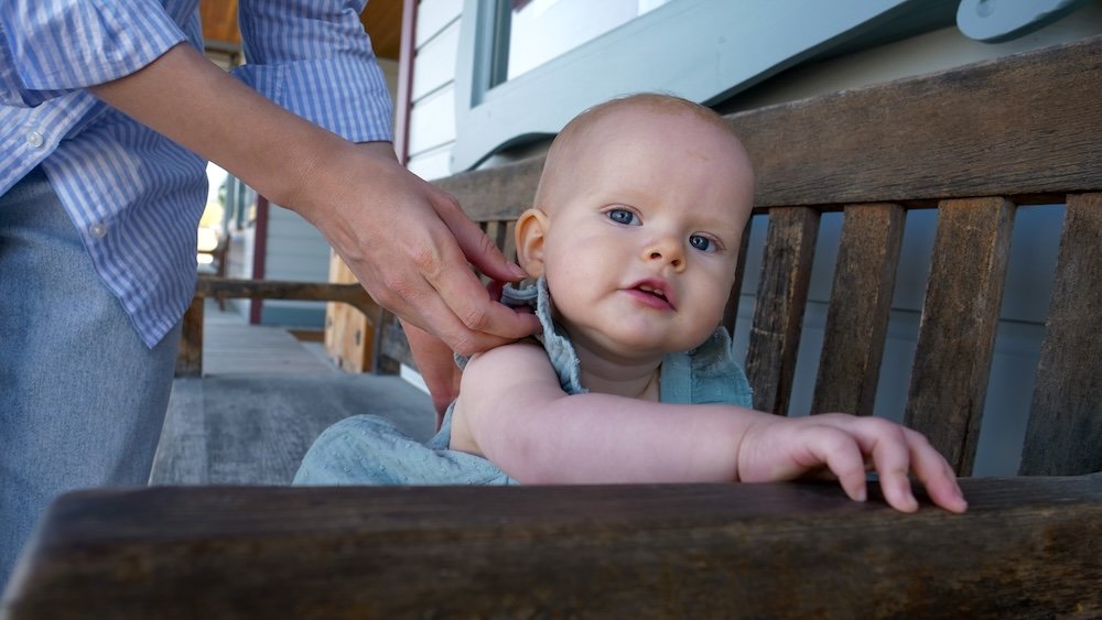Baby Aurelia on a bench at Fort Steele At Fort Steele near Cranbrook, British Columbia, baby Aurelia sits on a wooden bench while Audrey Bergner of That Backpacker adjusts her dress, with heritage-style buildings and porch details creating a warm, historic backdrop for the moment.
