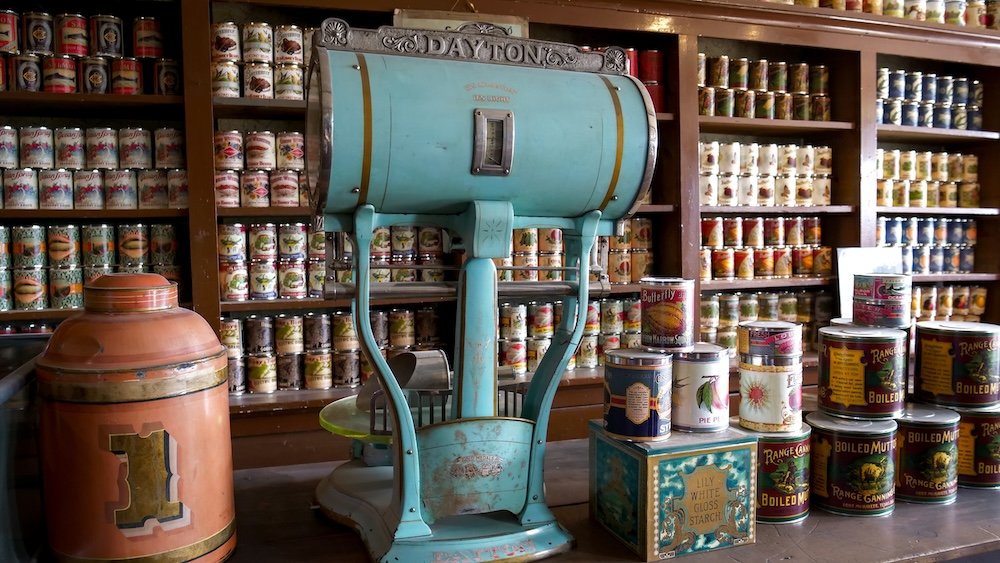 Historic Dayton scale surrounded by shelves of vintage canned goods on display at Fort Steele Heritage Town near Cranbrook, BC, illustrating how food was weighed, stored, and sold in general stores during the frontier and boomtown era.