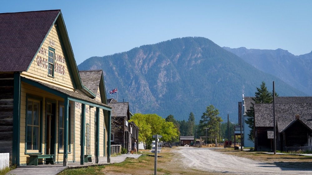 Fort Steele Heritage Town near Cranbrook, BC, with historic wooden buildings lining the dirt road and dramatic Rocky Mountain foothills rising behind them, showing the preserved boomtown streetscape and scenic Kootenay setting visitors experience today.
