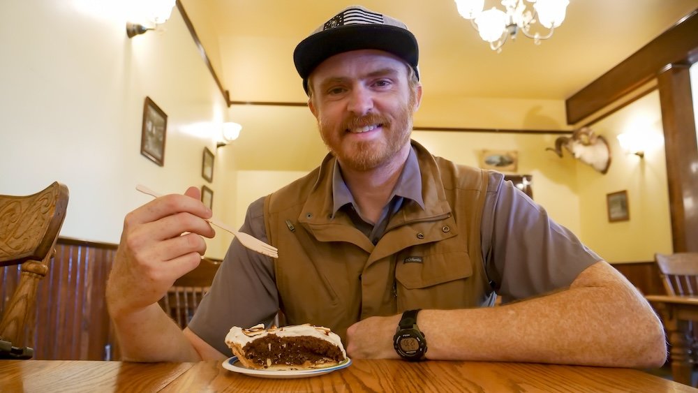 Fort Steele Heritage Town near Cranbrook, BC, inside a historic dining room where Nomadic Samuel enjoys a slice of rich peanut butter pie, highlighting the old-fashioned desserts and food stops that make a family day trip feel cozy and memorable.