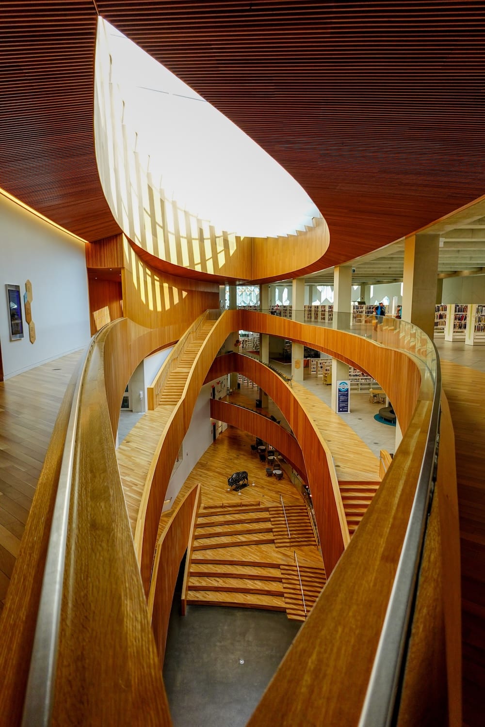 Una vista en picado a través del atrio de madera de cuatro niveles de la Biblioteca Central de Calgary en Calgary, Alberta, Canadá, mostrando escaleras curvas, estantes de libros y la pequeña escultura de un bisonte visible en la planta baja abajo.
