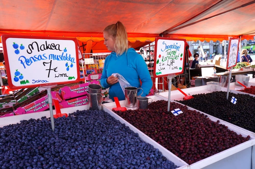 Fresh Finnish berries displayed in large trays at an outdoor Helsinki market stall, including blueberries and several darker seasonal varieties sold by the litre during summer in Finland.
